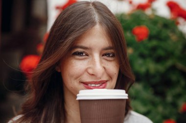 Kahve fincanı tutarak ve şehirde arka planını çiçekler üzerinde sokak gülümseyen güzel saçlı şık hipster kız. Kafe Teras vaktinizi muhteşem mutlu genç kadın