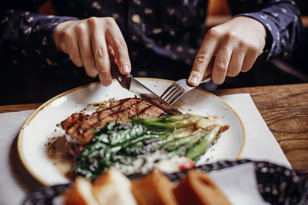 Hands holding fork and knife and eating delicious juicy steak with grilled cabbage,tomatoes and cheese on table at cafe in city street. Man tasting bbq with vegetables in restaurant