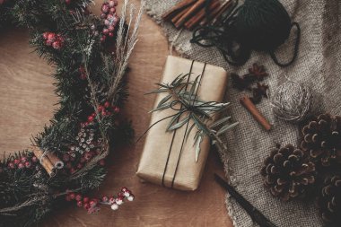 Rustik Noel çelenk ve şık zanaat hediye kutusu ile yeşil şubesine köknar, kırmızı meyveler, çam kozalakları, ip, tarçın, rustik ahşap, düz için makaslar yatıyordu. Atmosferik moody görüntü