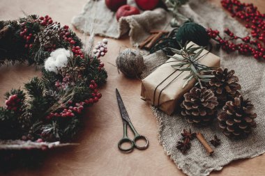 Rustik Noel çelenk ve şık zanaat hediye kutusu, çam dalları ile kırmızı meyveler, çam kozalakları, ip, tarçın, makas rustik ahşap üzerine. Kış tatil Atölyesi'nde atmosferik moody görüntü