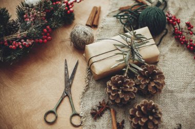 Rustik Noel çelenk ayrıntıları. Çam dalları kırmızı meyveler, çam kozalakları, rustik ahşap üzerine tarçın, makas ve zanaat hediye kutusu ile. Kış tatil Atölyesi'nde atmosferik moody görüntü