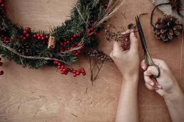 Holding eller, rustik Noel çelenk yapım makas otlar, çam dalları, kırmızı meyveler, çam kozalakları rustik ahşap zemin üzerinde. Kış tatil Atölyesi'nde atmosferik moody görüntü