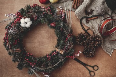 Rustik Noel çelenk, düz yatıyordu. Çam dalları, elma rustik ahşap arka plan üzerinde kırmızı meyveler, çam kozalakları, şerit, makas, tarçın, pamuk ile. Atmosferik moody görüntü