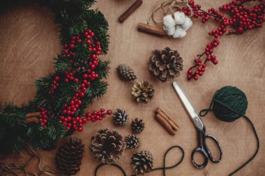 Rustik Noel çelenk düz yatıyordu. Çam dalları, çelenk, kırmızı meyveler, çam kozalakları, iplik, makas, tarçın, pamuk rustik ahşap zemin üzerinde. Noel çelenk, yapma