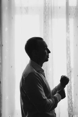 Şık damat beyaz gömlek koyarak pencere düğün töreni önce yumuşak ışık sabah hazırlanıyor