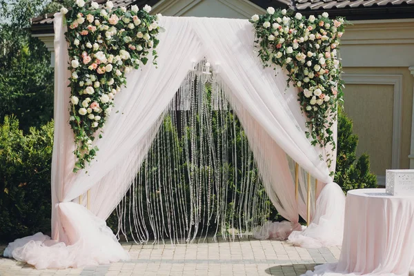 Stylish modern wedding arch with roses,tulle,stones, floral decorations at wedding reception outdoors. Luxury adorning.