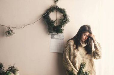 Örme kazak el yapımı Noel çelenk ve ışıklar arka plan üzerinde rustik odasında poz şık hippi kız. Atmosferik rahat anlar, kış tatili. Moody görüntü