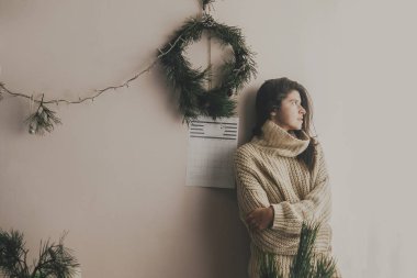 Örme kazak el yapımı Noel çelenk ve ışıklar arka plan üzerinde rustik odasında poz şık hippi kız. Atmosferik rahat anlar, kış tatili. Moody görüntü