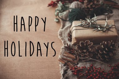 Mutlu tatil metin şık rustik Noel hediye kutusu çam dalları, kırmızı meyveler, çam kozalakları, rustik ahşap üzerine tarçın ile oturum. Atmosferik görüntü. Mevsimlik tebrik kartı