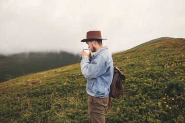 Çay içme ve üstünde tepe-in arka plan sisli dağların tepelerinde daimi şık hipster seyahat. Uzay kopyalayın. Yakışıklı adam dağlarında seyahat sırt çantası ile şapkalı. Yolculuk tutkusu