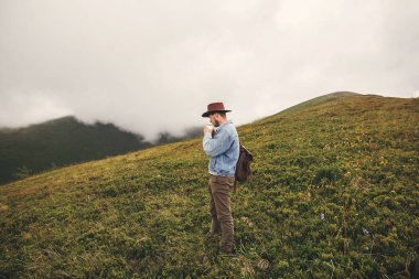 Çay içme ve üstünde tepe-in arka plan sisli dağların tepelerinde daimi şık hipster seyahat. Uzay kopyalayın. Yakışıklı adam dağlarında seyahat sırt çantası ile şapkalı. Yolculuk tutkusu