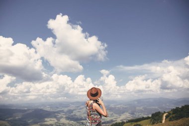 Stylish hipster girl in hat walking on top of sunny mountains. Happy young woman exploring in summer mountains and looking at sky. Travel and wanderlust concept. clipart