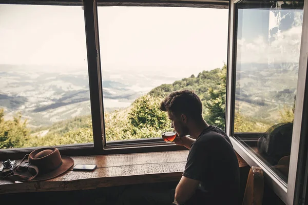 时尚的嬉皮士旅客在玻璃杯中喝着热茶 背景是美丽的树林和山脉 从山上的木窗可以看到风景 文本的空间 — 图库照片