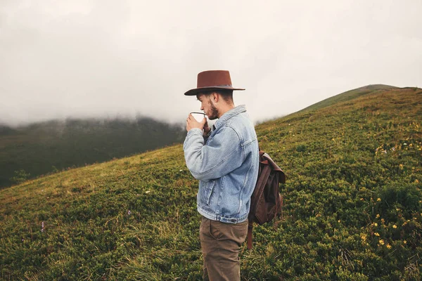 时尚的嬉皮士旅客喝茶 站在山顶上的雾蒙蒙的山 复制空间 英俊的男人戴帽子背包在山上旅行 流浪欲望 — 图库照片
