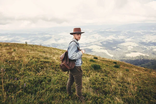 时尚的嬉皮士旅客喝茶 站在山顶上的雾蒙蒙的山 复制空间 英俊的男人戴帽子背包在山上旅行 流浪欲望 — 图库照片