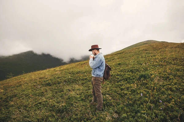 时尚的嬉皮士旅客喝茶 站在山顶上的雾蒙蒙的山 复制空间 英俊的男人戴帽子背包在山上旅行 流浪欲望 — 图库照片