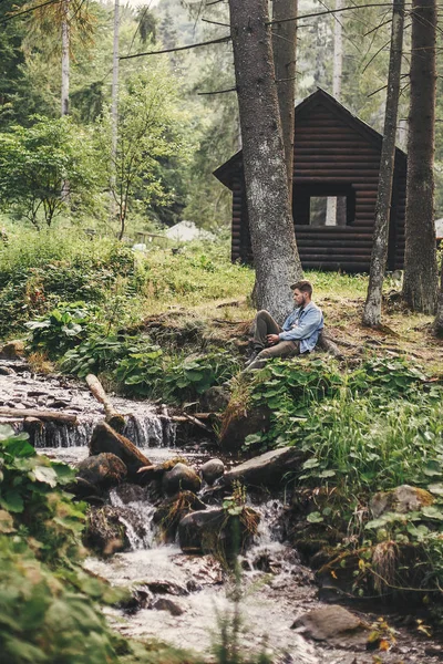 时尚的嬉皮士旅行者坐在森林里的河流背景下 在山上的木木 探索夏天的树林 复制空间 旅行和流浪欲望的概念 与自然放松 — 图库照片