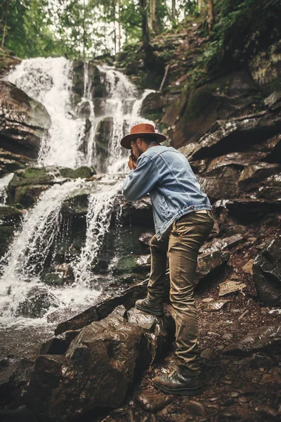穿着帽子在山上旅行的时尚年轻人 希普斯特男子与相机拍摄美丽的瀑布照片 探索夏季森林 旅行和流浪欲望的概念 文本的空间 — 图库照片