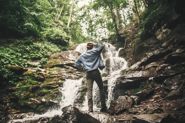 旅行和流浪欲望的概念 穿着帽子在山上旅行的时尚年轻人 希普斯特男子看着美丽的瀑布在夏季森林 文本的空间 — 图库照片