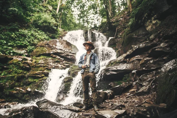旅行和流浪欲望的概念 穿着帽子在山上旅行的时尚年轻人 希普斯特男子与照片相机探索美丽的瀑布在夏季森林 文本的空间 — 图库照片