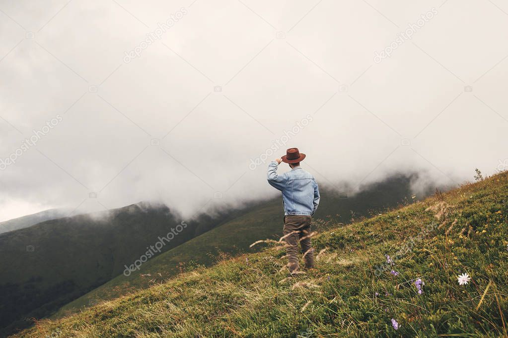 Stylish hipster traveler in hat standing on top of hills on  background of foggy mountains. Copy space. Travel and wanderlust concept.