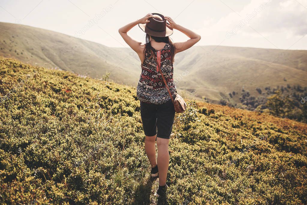 Stylish hipster girl in hat traveling on top of sunny mountains. Young beautiful woman with beautiful hair traveling and exploring mountains in light. Travel and wanderlust concept