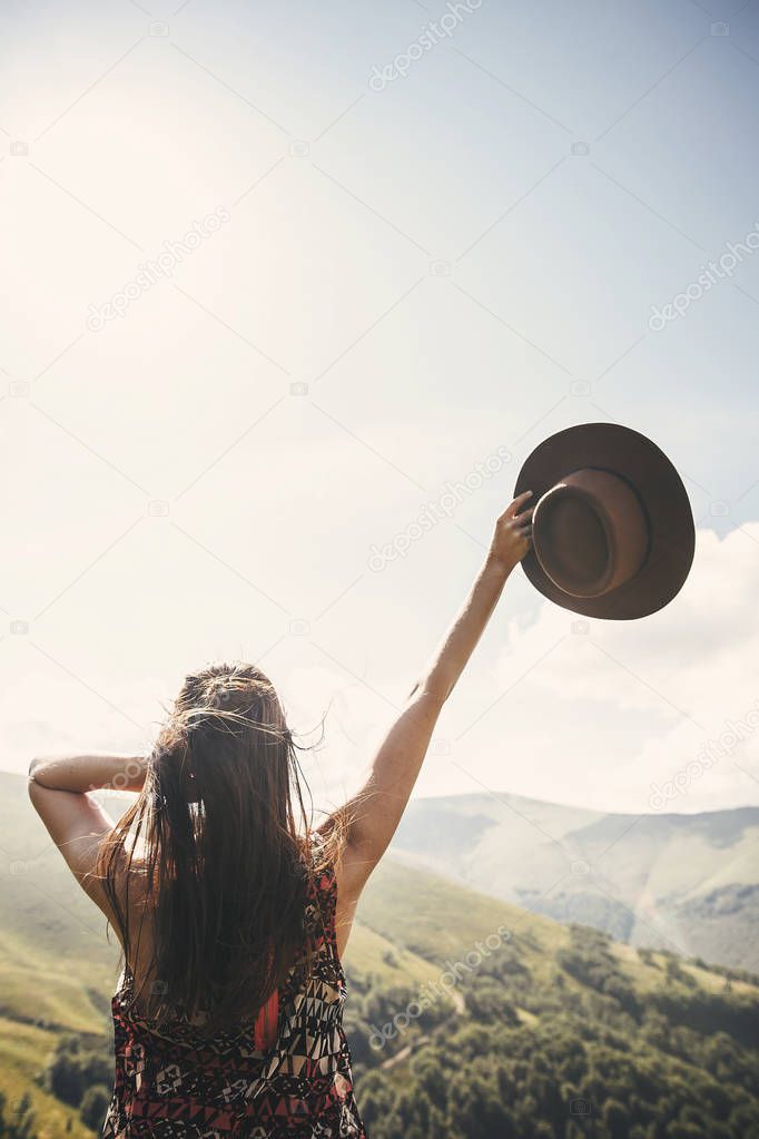 Stylish hipster girl in hat traveling on top of sunny mountains. Young beautiful woman with beautiful hair exploring mountains  and walking in light. Travel and wanderlust concept.