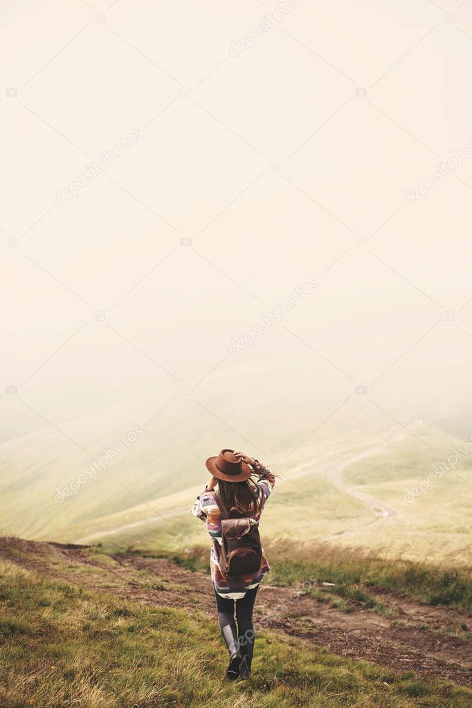 Stylish hipster girl in hat walking on top of mountains. Happy young woman with backpack exploring misty mountains. Travel and wanderlust concept. Amazing atmospheric moment
