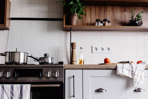 Приготовление пищи на современной кухне со стальной духовкой, кастрюлями, ножом на деревянной разделочной доске с овощами, перцем, специями, маслом на деревянной столешнице. Домашняя еда. Стильная кухонная мебель серого цвета
