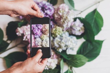 Telefon ve ortanca çiçek alarak fotoğraf üzerinde rustik ahşap beyaz holding eller, düz yatıyordu. İçerik Sosyal medya kavramı, bloglama fotoğraflar için. Mutlu anneler günü. Metin için yer