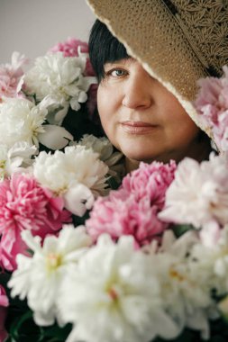 Birçok şakayık çiçekleri ile poz şapkalı güzel bir kadın. Büyük buket pembe peonies çocuklardan mutlu anne. Mutlu anneler günü kavramı. Uluslararası Kadınlar günü