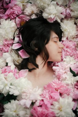 Portrait of boho woman with natural makeup lying in peonies. Creative floral photo. Aroma and spa concept. International Womens Day. Beautiful brunette girl in many pink and white flowers