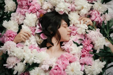 Portrait of boho woman with natural makeup lying in peonies. Creative floral photo. Aroma and spa concept. International Womens Day. Beautiful brunette girl in many pink and white flowers
