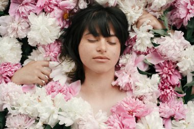 Portrait of boho woman with natural makeup lying in peonies. Creative floral photo. Aroma and spa concept. International Womens Day. Beautiful brunette girl in many pink and white flowers