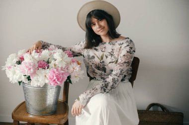 Büyük peonies buket ile poz şapkalı güzel esmer boho kız. Metal kova ile peonies rustik ahşap sandalye üzerinde oturan şık hipster kadın. Uluslararası Kadınlar günü