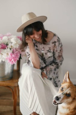 Stylish boho girl playing and smiling with cute golden dog at metal bucket with peonies on rustic wooden chair in home. Beautiful hipster woman having fun with her doggy. Friendship