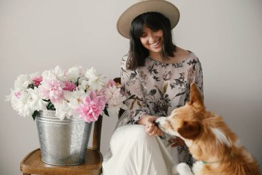 Metal kova ile peonies rustik ahşap sandalye üzerinde oturan ve sevimli golden köpekle oynamayı şık boho kız. Onu evde beslenen hayvan vasıl büyük peonies buket ile oynarken güzel hipster kadın.