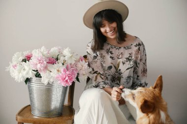 Stylish boho girl playing and smiling with cute golden dog at metal bucket with peonies on rustic wooden chair in home. Beautiful hipster woman having fun with her doggy. Friendship