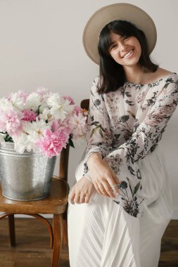 Şapkalı büyük peonies buket ile gülümseyen güzel esmer boho kız. Metal kova ile peonies rustik ahşap sandalye üzerinde oturan şık hipster kadın. Uluslararası Kadınlar günü