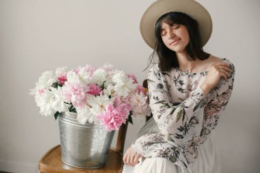 Metal kova ile peonies rustik ahşap sandalye üzerinde oturan şapkalı şık boho kız. Güzel hipster kadın büyük peonies buket ile poz. Kırsal yaşam. Uluslararası Kadınlar günü
