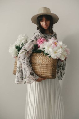 Blurred defocused image of boho girl holding pink and white peonies in rustic basket. Stylish hipster woman in hat and bohemian floral dress with flowers. International Womens Day