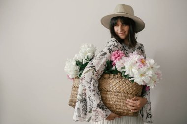 Pembe ve beyaz peonies rustik sepet içinde tutarak boho kız şehvetli portresi. Şapka ve çiçeklerle poz bohem çiçek elbise şık hipster kadın. Uluslararası Kadınlar günü.