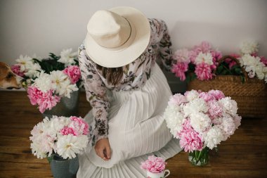Boho kız köpeğini, pembe ve beyaz peonies ile rustik ahşap yere oturma. Şapka ve çiçeklerle bohem çiçek elbise şık hipster kadın. Kırsal yaşam