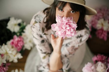 Pembe ve beyaz peonies rustik sepeti ve ahşap zemin üzerinde metal kova içinde oturan Boho kız. Pembe şakayık çiçekleri arasında tutarak bohem içinde şık hipster kadın. Kırsal yaşam