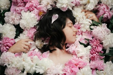 Portrait of boho woman with natural makeup lying in peonies. Creative floral photo. Aroma and spa concept. International Womens Day. Beautiful brunette girl in many pink and white flowers