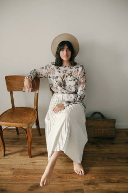 Güzel esmer boho kız evinde rustik ahşap sandalye üzerinde oturan şapkalı. Bohem elbiseyle poz şık hipster kadın. Uluslararası Kadınlar günü. Kırsal yaşam