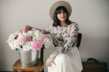 Metal kova ile peonies rustik ahşap sandalye üzerinde oturan şapkalı şık boho kız. Güzel hipster kadın büyük peonies buket ile poz. Kırsal yaşam. Uluslararası Kadınlar günü