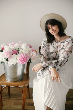 Metal kova ile peonies rustik ahşap sandalye üzerinde oturan şapkalı şık boho kız. Güzel hipster kadın büyük peonies buket ile poz. Kırsal yaşam. Uluslararası Kadınlar günü