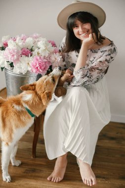 Stylish boho girl playing and smiling with cute golden dog at metal bucket with peonies on rustic wooden chair in home. Beautiful hipster woman having fun with her doggy. Friendship