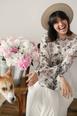 Metal kova ile peonies rustik ahşap sandalye üzerinde oturan ve sevimli golden köpekle oynamayı şık boho kız. Onu evde beslenen hayvan vasıl büyük peonies buket ile oynarken güzel hipster kadın.